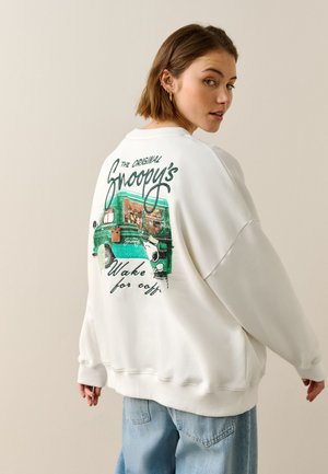 Ung kvinde iført hvid sweatshirt med Snoopy-grafik og grøn vogn, der kigger over sin skulder mod en ensfarvet beige baggrund.