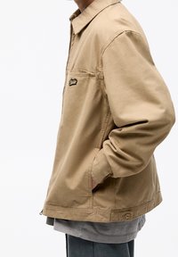Chaqueta de algodón beige con cremallera frontal y bolsillos laterales. Presenta un parche negro con el logo en el pecho, con un diseño de corte relajado.