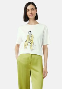 Weißes T-Shirt mit kurzen Ärmeln, das eine illustrierte Figur in Gelb und Schwarz zeigt, begleitet von dem Text "Fortsetzung folgt...", kombiniert mit glänzenden, grünen weit geschnittenen Hosen.