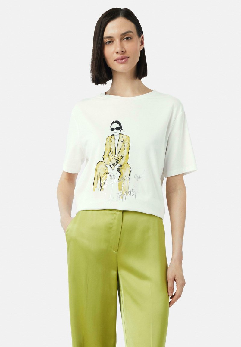 Weißes T-Shirt mit kurzen Ärmeln, das eine illustrierte Figur in Gelb und Schwarz zeigt, begleitet von dem Text "Fortsetzung folgt...", kombiniert mit glänzenden, grünen weit geschnittenen Hosen.