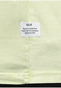 Lichtgroene stof met een gestructureerd oppervlak. Een wit kledinglabel met zwarte tekst dat merk- en verzorgingsinformatie vermeldt is zichtbaar.