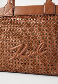 La borsa a tracolla intrecciata marrone presenta una base testurizzata simile alla pelle. Un dettaglio notevole include una firma prominente e liscia dello stesso colore.