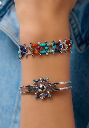 Two bracelets on a wrist: a multicolored beaded bracelet with silver accents and a silver cuff bracelet with a floral design and colored gems.