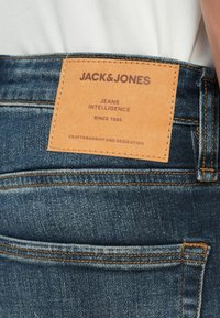 Zilnas džinsi ar gaiši zilu krāsu, ar oranžām šuvēm un brūnu ādas siksniņu, uz kuras ir uzraksts "JACK & JONES" jostas daļā.