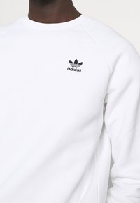 Vit bomullströja med en svart Adidas-logotyp på vänster bröst, med rund halsringning och kontrasterande raglanärmar.
