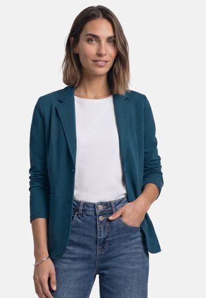 Kvinde med skulderlangt brunt hår iført en teal blazer over en hvid skjorte og blå jeans, den ene hånd i lommen, smilende let.