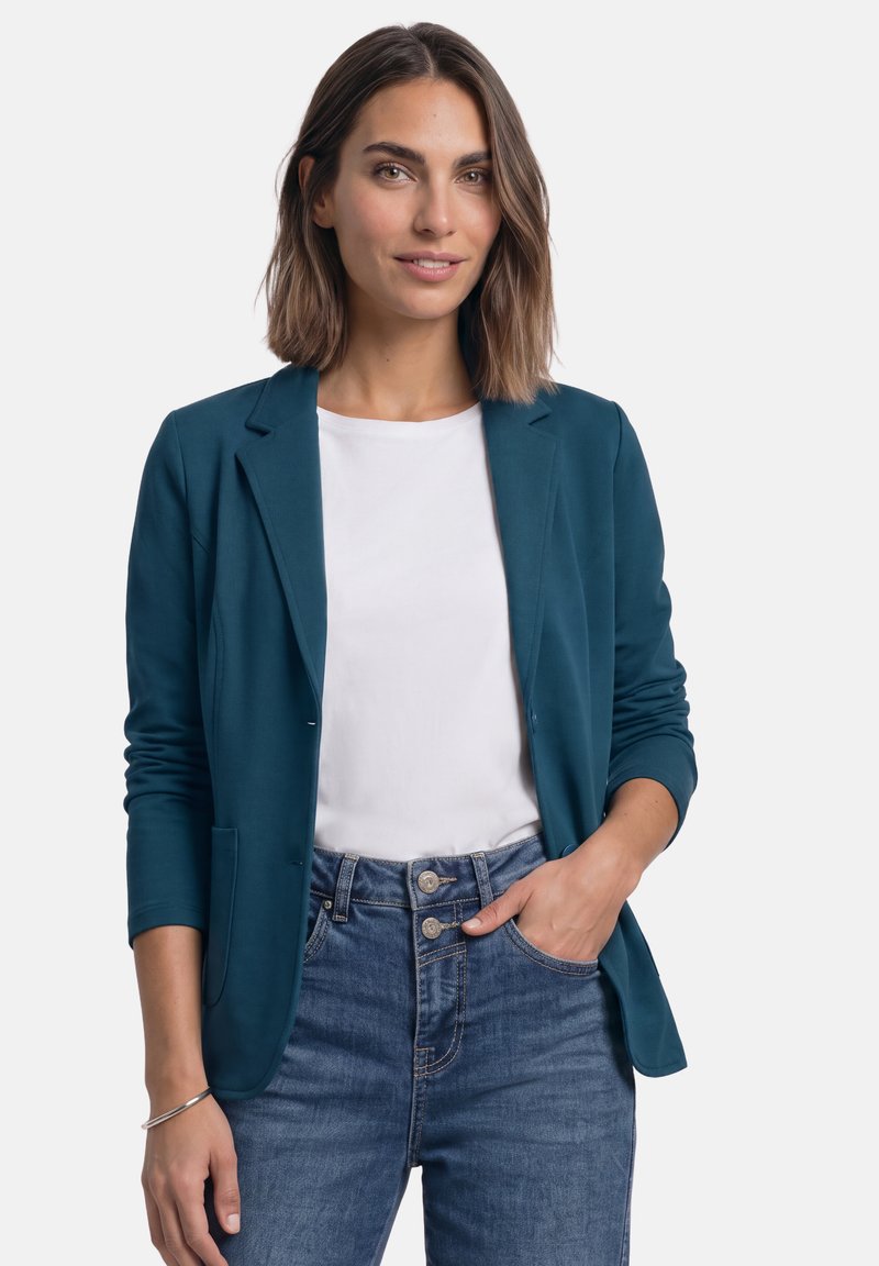 Femme aux cheveux châtain mi-longs portant un blazer bleu sarcelle sur une chemise blanche et un jean bleu, une main dans la poche, souriant légèrement.