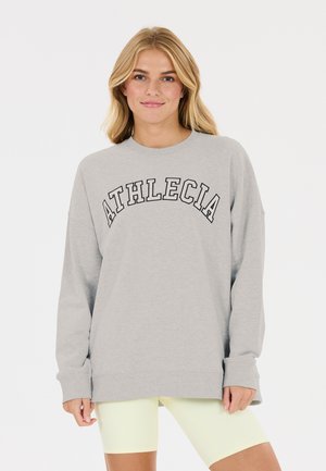 Blonde Frau trägt ein hellgraues Oversize-Sweatshirt mit dem Schriftzug "ATHLECIA" und blassgelbe Radlerhosen, steht vor weißem Hintergrund.
