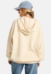Crème-kleurige oversized hoodie met een gestructureerde fleece binnenkant en een verstelbare trekkoordkap, gecombineerd met een lichtblauwe denim pet en jeans.