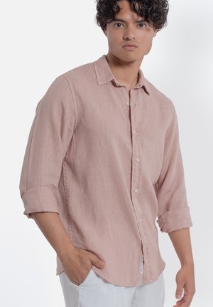 Giovane uomo con capelli ricci che indossa una camicia rosa chiaro con bottoni e maniche arrotolate e pantaloni bianchi, in piedi con una mano in tasca.