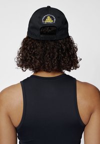 Schwarze Baseballkappe mit gesticktem Design, das eine gelbe Grafik zeigt. Verstellbarer Riemen auf der Rückseite. Getragen mit einem ärmellosen schwarzen Oberteil.
