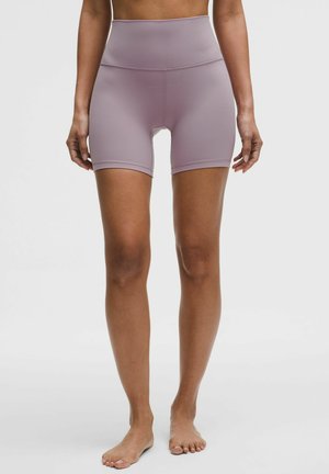 Vrouw met hoge taille mauve sportieve fietsshorts, staand op blote voeten tegen een lichtgrijze achtergrond.