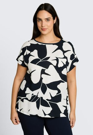 Femme portant un haut noir à manches courtes avec de grands motifs abstraits de feuilles blanches, debout devant un fond clair uni.