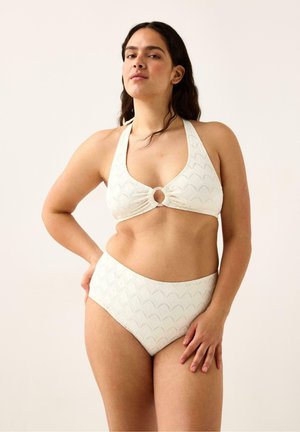 Kvinde iført hvid tekstureret halterbikini med cirkulær ringdetalje ved brystet, stående med den ene hånd på hoften mod en ensfarvet baggrund.