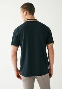 Mörk marinblå polo med kort ärm och en texturerad krage med beige ränder. Modellen står med ryggen mot kameran, iförd ljusa byxor.