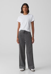 T-shirt en coton blanc et pantalon large en velours côtelé gris avec taille élastique et cordon de serrage. Chaussures blanches avec une semelle claire.