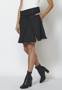 Femme portant une jupe noire texturée aux bords effilochés et des bottines noires à talons sur un fond uni.
