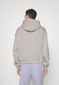 Homme portant un sweat à capuche gris clair et un pantalon lavande, debout de dos devant un fond uni.