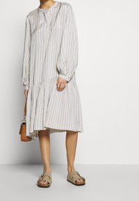 Lehký pruhovaný šaty s dlouhými nafouknutými rukávy, sklady na spodním lemu a zapínáním na knoflíky, doplněné béžovými sandály a hnědou taškou.