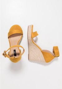 Žluté semišové klínové sandály s pletenou jutovou podrážkou, otevřenou špičkou a páskem kolem kotníku. Mají hladký interiér a minimalistický design.