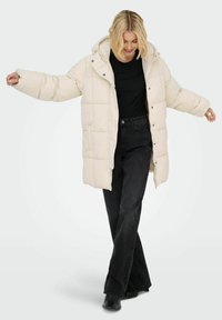 Abrigo acolchado de color crema con capucha y cierres de botón. Presenta un diseño acolchado, corte holgado y se lleva sobre una camisa negra y jeans.