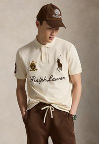 Νέος άνδρας που φοράει μπεζ μπλούζα πόλο Ralph Lauren με κεντημένα λογότυπα, καφέ καπέλο και καφέ παντελόνι με κορδόνι, χέρια στις τσέπες, κοιτάζοντας προς τα κάτω.