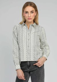 Chemise à boutons avec un motif rayé vertical blanc et noir, dotée d'un col, de manches longues et de détails décoratifs le long des bords.