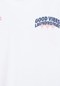 Tissu blanc avec texte bleu marine "GOOD VIBES LAUNDROMAT" et trois icônes d’étoile rouge sous le texte en haut à droite.