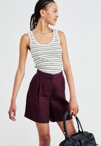 Stripet tank topp i krem og marinblå, kombinert med burgunder høyt liv-shorts med knappefeste. Modellen holder en svart håndveske.