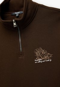 Jersey marrón con un cuello de media cremallera, que presenta gráficos de montañas y el texto "Las montañas llaman y debo ir". Textura suave.