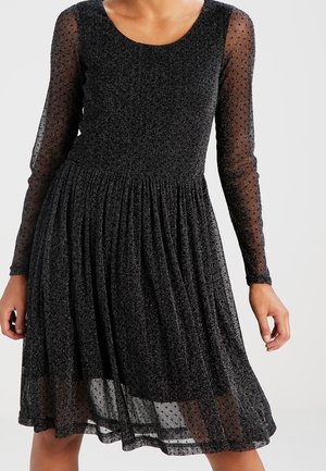 Kobieta w czarnej sukience sięgającej do kolan, z długimi rękawami z przezroczystym materiałem w kropki oraz błyszczącą, plisowaną spódnicą.