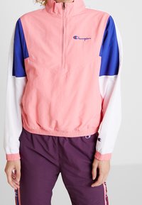 Rompevientos ligero en rosa, azul y blanco. Cuenta con media cremallera, dobladillo elástico y detalle del logo. Incluye mangas largas con puños elásticos.