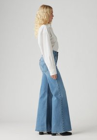 Világoskék, bő szárú, magas derekú farmernadrág. Fehér, hosszú ujjú, gombos inggel és fekete, magas sarkú csizmával párosítva.