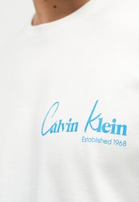 Λευκό πουκάμισο με ανοιχτό γαλάζιο λογότυπο "Calvin Klein Established 1968" στο στήθος, φορεμένο από άτομο με μέτριο τόνο δέρματος.