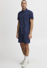 Granatowa koszula na krótki rękaw zapinana na guziki z kieszenią na piersi oraz pasujące granatowe szorty. Tkanina ma fakturowane wykończenie. Noszone z białymi trampkami.