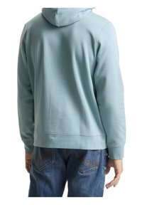 Personne portant un sweat à capuche bleu clair et un jean bleu vue de dos, debout avec la main droite détendue sur le côté.