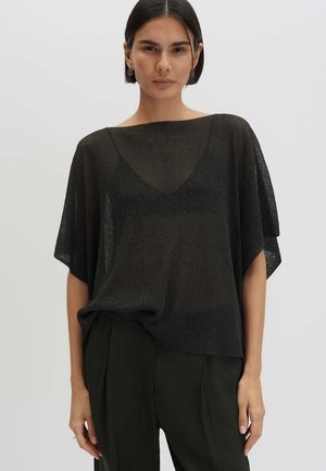 Femme portant un haut noir ample et transparent par-dessus un soutien-gorge noir, associé à un pantalon noir taille haute sur un fond clair uni.