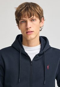 Sudadera con cremallera en color azul marino, fabricada en un tejido suave, con capucha ajustable y un pequeño logotipo rojo en el lado izquierdo del pecho. Llevada sobre una camiseta blanca.