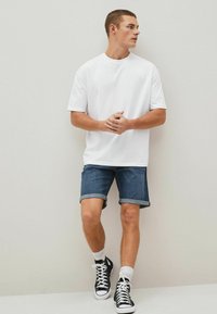 Valge lühikeste varrukatega T-särk, denim-lühikesed püksid, mille ääred on üles keeratud, mustad kõrge säärega spordijalatsid ja valged sokid. Modell seisab ühtlase tausta ees.