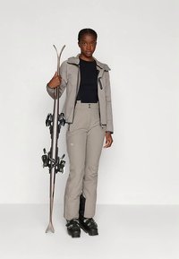 Mlada žena u bež skijaškom odijelu i crnim čizmama drži par skija, stojeći nasuprot običnoj svijetloj pozadini.