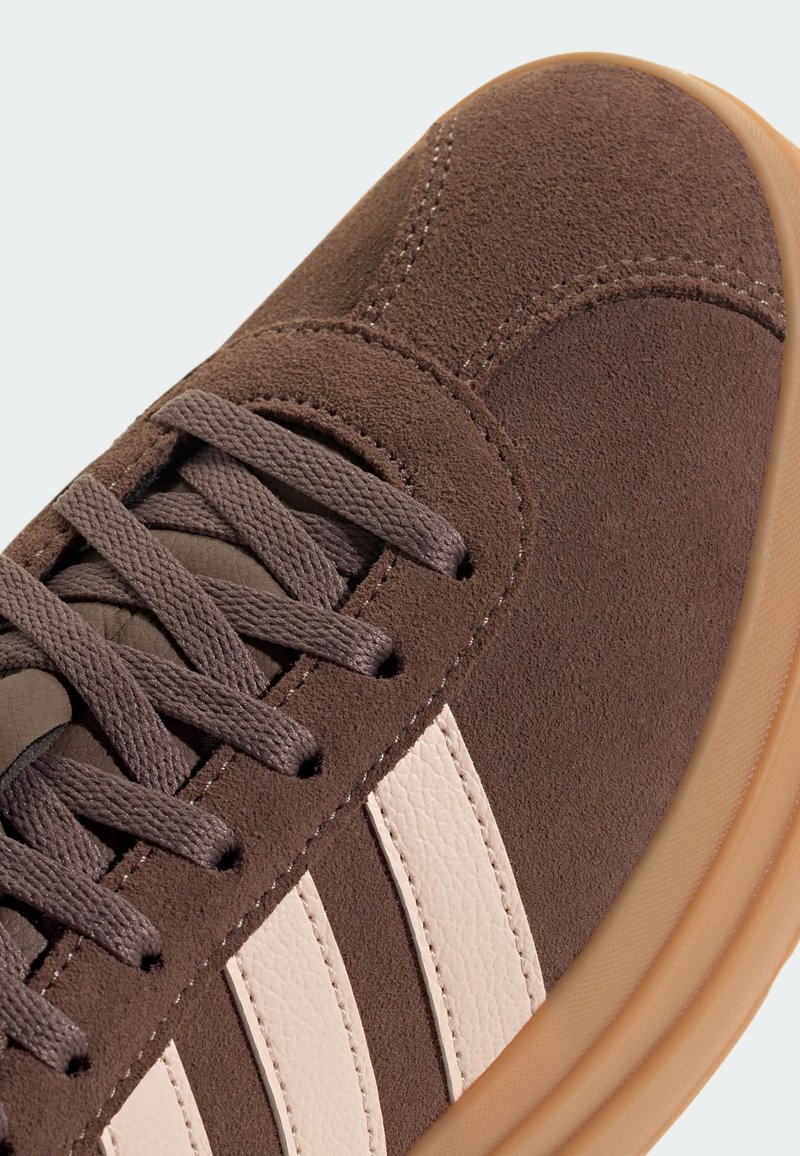 Bruine suède sneakers met drie witte leren strepen, gestructureerde bovenkant en bruine veters. Lichte tan rubberen zool met minimale details.