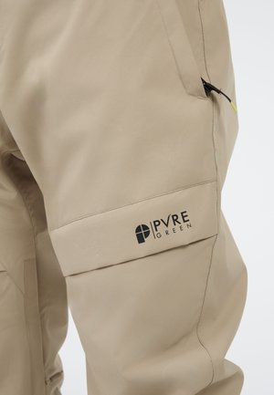 Beige waterdichte broek met een gladde textuur, voorzien van een ritszak en een zwart logo "PVRE GREEN" op het bovenbeen.