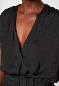 Blusa nera con scollatura a V profonda e maniche corte. Presenta una texture morbida e liscia e un dettaglio arricciato in vita.