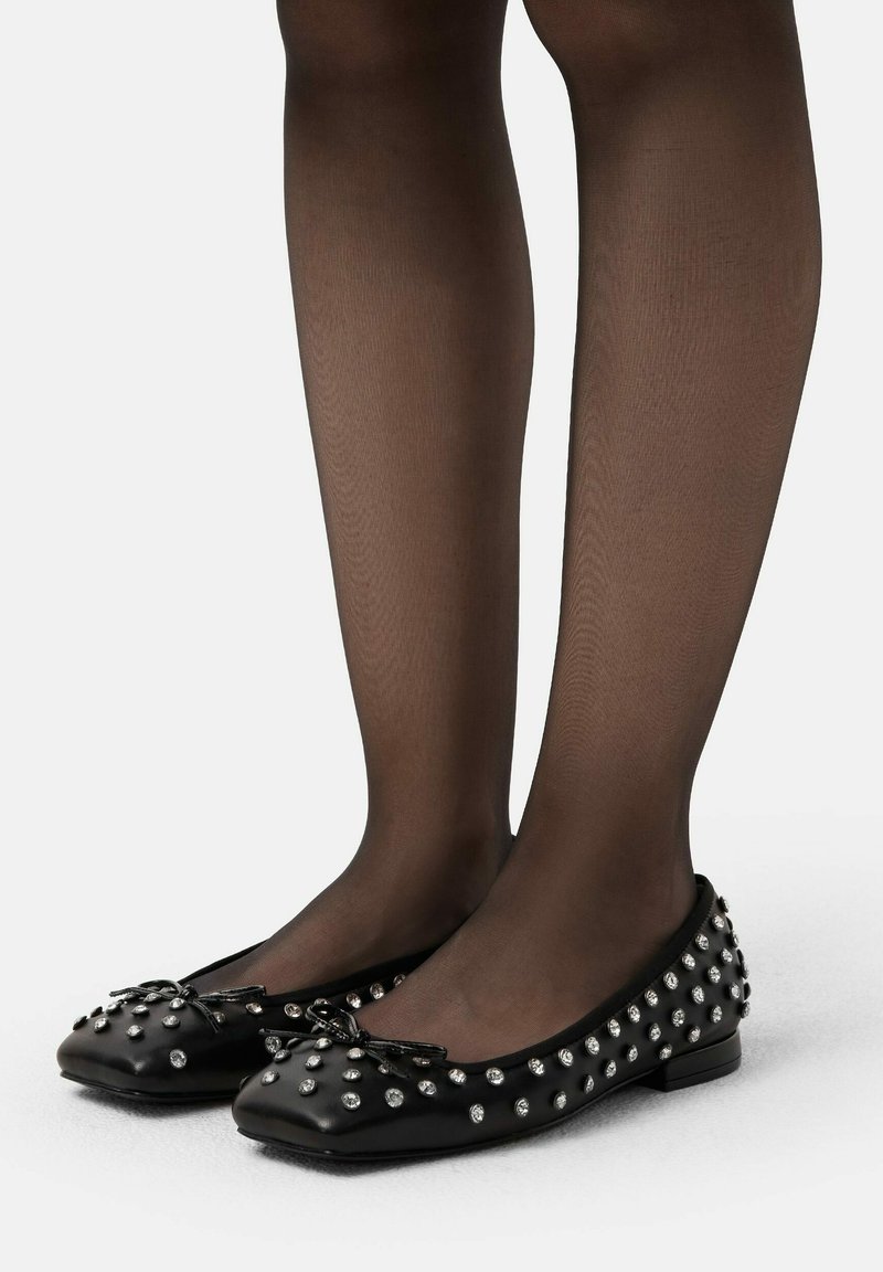 Beine, die durchsichtige schwarze Strumpfhosen und schwarze Ballettschuhe mit kleinen silbernen Nieten und einer Schleife oben tragen, stehen auf einem weißen Boden.