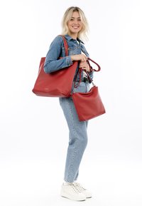 Sac en cuir rouge avec une texture granuleuse et des accessoires en métal argenté, tenu à côté d'un petit sac assorti. Tenue décontractée en denim et baskets.