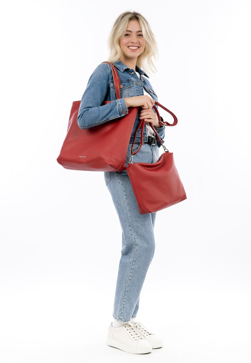 Sac en cuir rouge avec une texture granuleuse et des accessoires en métal argenté, tenu à côté d'un petit sac assorti. Tenue décontractée en denim et baskets.