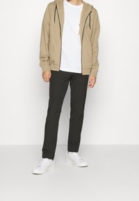 Beige Kapuzenjacke mit Reißverschluss und Kordelzug, getragen über einem weißen T-Shirt, kombiniert mit schwarzen Hosen und weißen Turnschuhen; lässiges, figurbetontes Design.