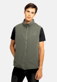 Veste sans manches verte avec col montant, fermeture éclair à l'avant et poches latérales, en matériau lisse et léger. Logo imprimé sur la poitrine.