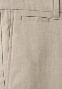 Gros plan sur un pantalon en lin beige avec une poche latérale, une poche passepoilée et une boucle de ceinture avec des détails visibles de couture.