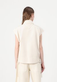 Top crema senza maniche realizzato in cotone. Caratterizzato da un colletto a punta e un orlo posteriore curvo. Dettagli di cuciture sottili sul colletto.